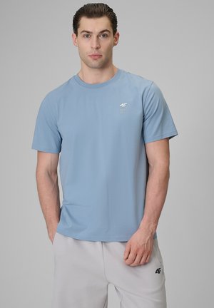 Młody mężczyzna z zaczesanymi do tyłu włosami nosi jasnoniebieski T-shirt z krótkim rękawem i jasnoszare szorty, stoi na jednolitym szarym tle.
