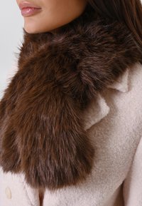Écharpe en faux fur marron avec une texture duveteuse, portée sur un manteau en laine rose clair avec des boutons ronds et un tissu doux et texturé.