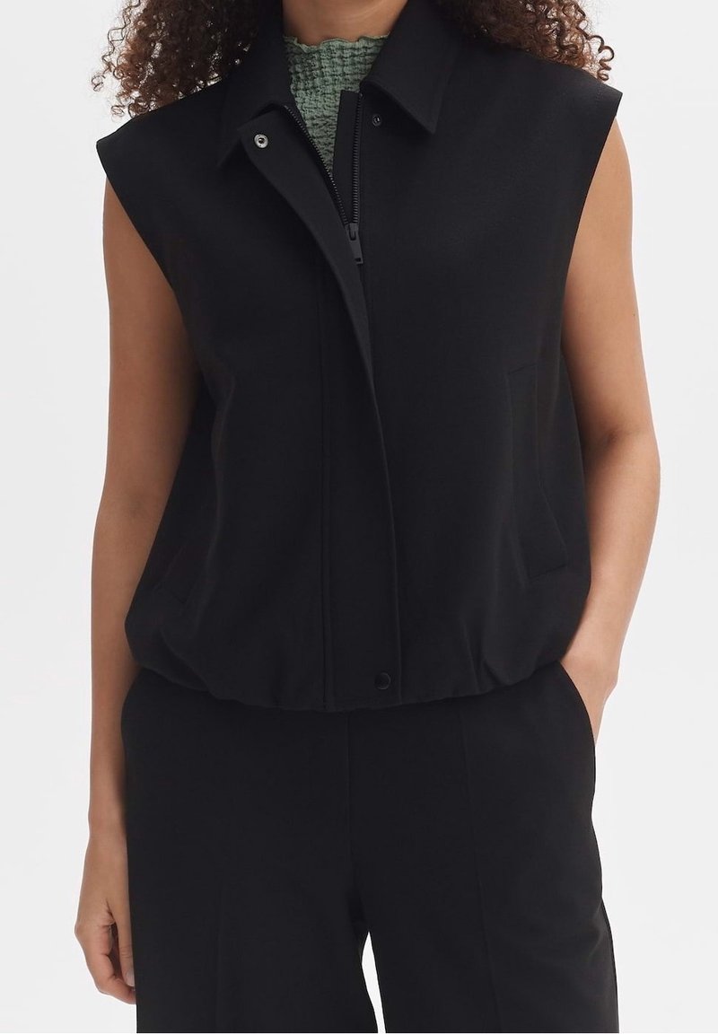 Czarna bezrękawnik z wysokim kołnierzem, z zamkiem z przodu i subtelnymi kieszeniami. Gładka tekstura, luźny krój i elastyczny detal u dołu.