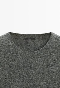 Szary wełniany sweter z okrągłym dekoltem. Tekstura jest miękka i puszysta, z delikatnym wzorem w różnych odcieniach szarości. Widoczna metka.