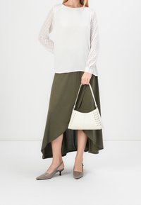 Blouse blanche avec manches transparentes à rayures, jupe asymétrique vert olive, talons gris, et sac blanc avec détails lacés.