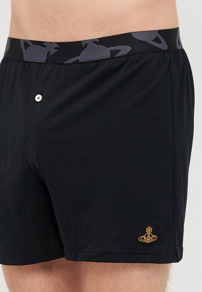 Schwarze Baumwoll-Boxershorts mit einem grauen gemusterten Bund. Sie verfügen über einen Knopfverschluss und ein kleines gelbes Logo an der unteren Seite.