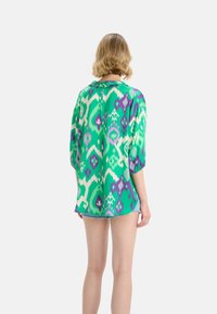 Kolorowa zielona bluzka z fioletowymi i białymi wzorami, charakteryzująca się luźnym krojem i krótkimi rękawami. Lekkie materiały o gładkiej fakturze.