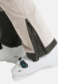 Beige skibroek met donkergroene accenten, voorzien van ritssluitingen aan de onderkant van de benen en een gladde textuur, gedragen over witte skibotten met blauwe details.