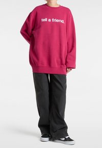 Personne portant un sweat-shirt rouge ample avec le texte « tell a friend. », un pantalon noir à jambes larges et des baskets noir et blanc, debout sur un fond blanc.