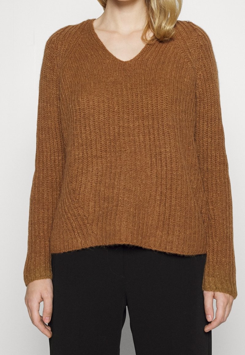 Pull en maille côtelée marron avec col en V porté par une personne, associé à un pantalon noir, raccourci au milieu du torse.