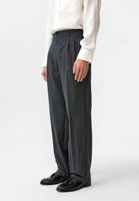Gestreepte grijze plooibroek met een hoge taille, voorzien van een knoopsluiting en zijzakken, gecombineerd met zwarte nette schoenen.