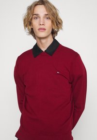 Sweater de malha vermelha com um colarinho preto, apresentando um pequeno logotipo do lado esquerdo. Mangas longas e punhos e bainha canelados.