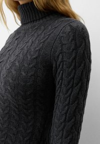 Pull en maille torsadée grise avec un col montant. Présente des motifs en relief et une finition texturée. Le tissu semble doux et chaud.