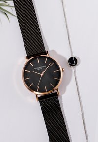 Reloj con correa de malla negra, caja redonda de oro rosa, esfera negra y marcadores en rosa. Pulsera plateada con un acento circular negro al lado.