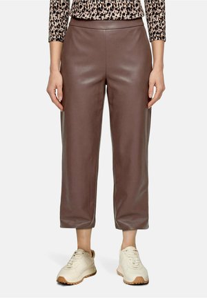 Femme portant un pantalon en cuir marron court, un haut à manches longues imprimé léopard et des baskets crème, debout avec les mains détendues.