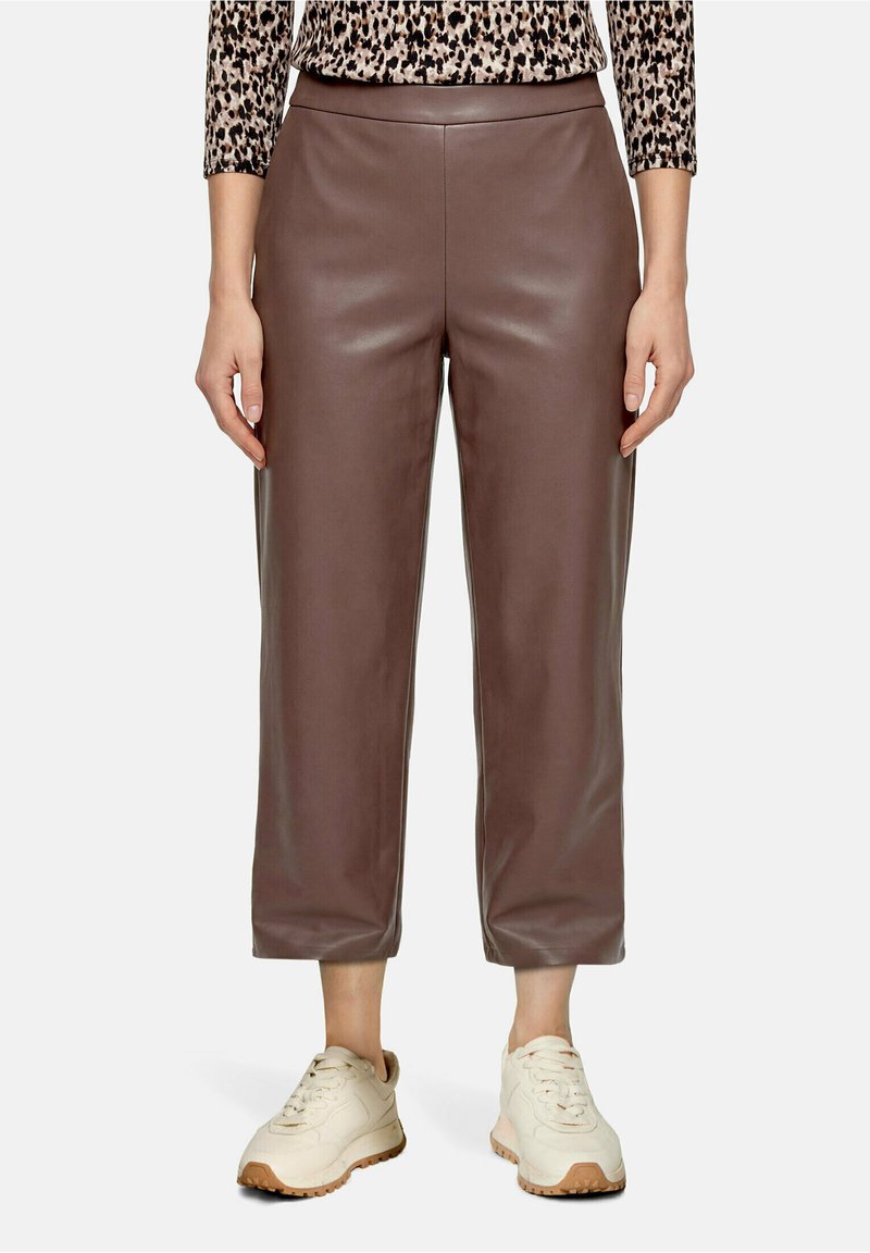 Femme portant un pantalon en cuir marron court, un haut à manches longues imprimé léopard et des baskets crème, debout avec les mains détendues.