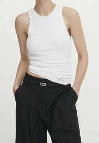 Hvid tætsiddende tanktop lavet af bomuld, kombineret med højtaljede sorte bukser med bælte. Toppen har rund halsudskæring og ærmeløs design.