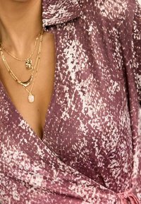 Abito a portafoglio in un motivo a pois mauve e bianco, caratterizzato da un profondo scollo a V, maniche corte e un dettaglio in vita annodato. Visibili più collane d'oro.