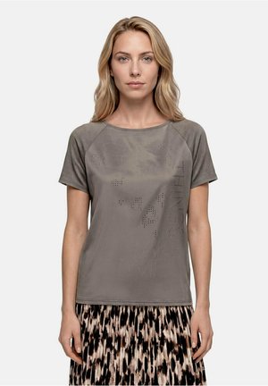 Femme blonde portant un t-shirt gris à manches courtes avec des détails textuels et une jupe noire et beige à motifs, debout devant un fond uni.