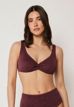 Mujer con cabello oscuro de longitud media que lleva un top de bikini morado oscuro con estampado y braguitas de talle alto sobre un fondo liso.