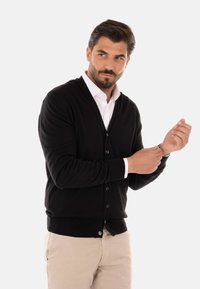 Cardigan nero realizzato in un tessuto morbido, con scollatura a V, chiusura a bottoni e polsini a coste, indossato sopra una camicia bianca e pantaloni beige.