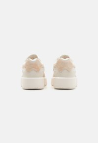 Las zapatillas cuentan con un upper de ante beige y cuero crema, con detalles en forma de onda, una suela texturizada y un diseño minimalista.