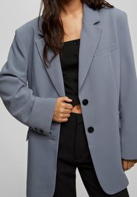 Blazer oversize gris clair avec un col cranté, deux boutons noirs et une poche poitrine unique, associé à un haut noir et un pantalon noir.