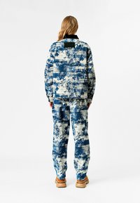 Conjunto de chaqueta y pantalones de corte relajado en tejido texturizado con un patrón abstracto en azul y crema. Presenta un color negro en el cuello y calzado marrón en contraste.