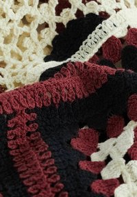 Hæklet tæppe med teksturerede mønstre i bordeaux, sort og creme. Distinkte bølgede kanter og cirkulære motiver gennem hele tæppet.