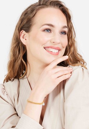 Gouden kettingarmband met elkaar verstrengelde schakels, gedragen om een pols, op een lichte beige stof. Gladde textuur, eenvoudig ontwerp, zonder versieringen.
