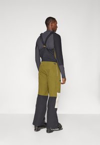 Calças de ski em bloco de cores verde oliva e azul marinho com alças, apresentando um ajuste ajustado, bolsos laterais e tecido texturizado. Sapatos pretos com logotipo.