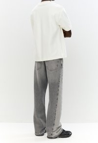 Weißes Baumwoll-T-Shirt und graue, ausgewaschene Jeans mit weitem Bein und markanten Gesäßtaschen, getragen mit schwarzen Sportschuhen.