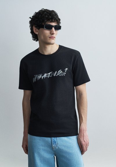 Young man with curly hair wearing black sunglasses, a black graphic t-shirt with distorted text, and light blue jeans, standing against a plain wall.