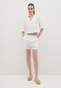 Witte geknipte shirt met korte mouwen en een decoratieve zak; gecombineerd met witte shorts met elastische taille en beige espadrilles.