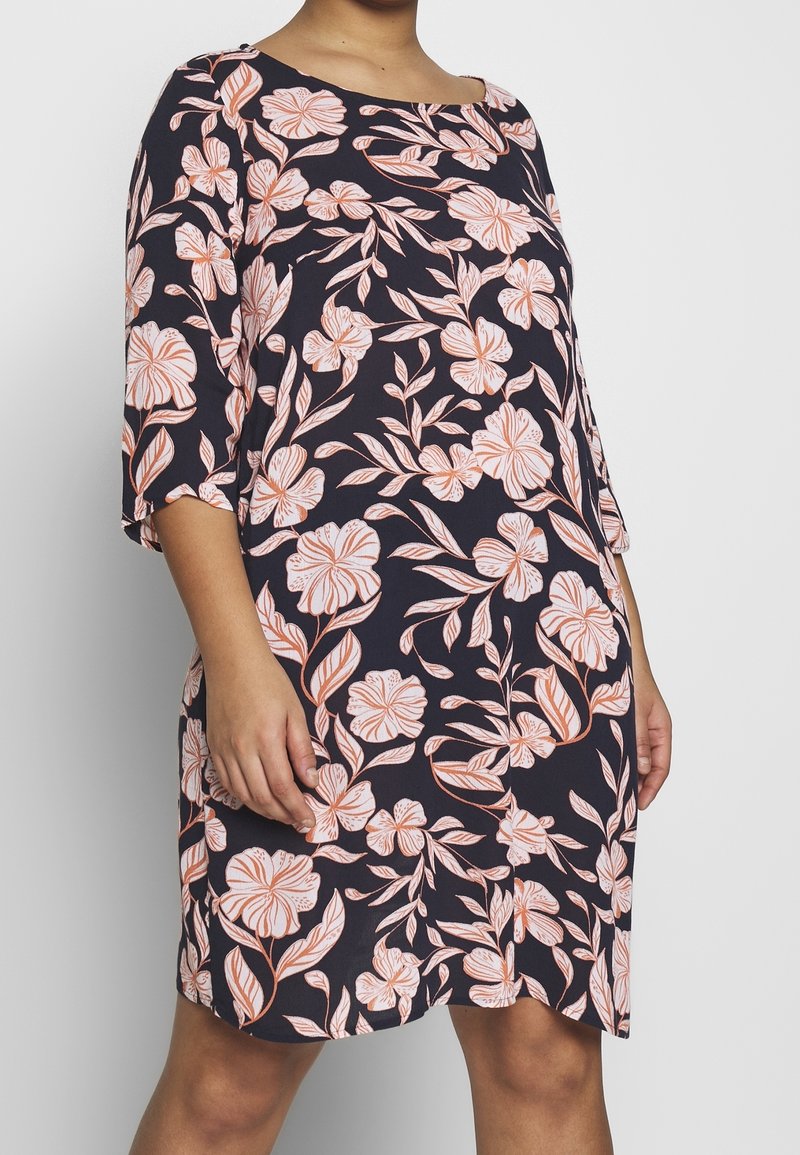 Femme portant une robe sombre avec des motifs floraux pêche et des manches trois-quarts, debout devant un fond clair uni.