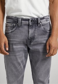 Jeans de denim gris con una textura ligeramente desgastada. Presentan cinco bolsillos, un cierre de botón y costuras contrastantes a lo largo de las costuras.