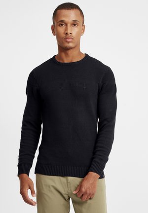 Maglione nero lavorato a maglia con una sezione superiore testurizzata, polsini e orlo a coste. Presenta un collo rotondo. Indossato con pantaloni chiari.