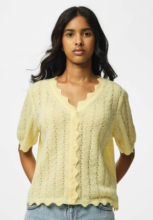 Jeune femme aux longs cheveux foncés portant un cardigan tricoté à manches courtes jaune pâle avec des bords festonnés et un jean bleu sur fond blanc.