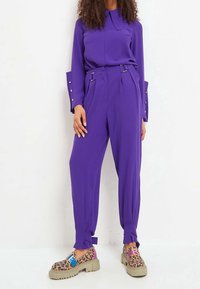 Blusa lunga manica viola con colletto; pantaloni a vita alta plissettati con passanti per cintura e laccetti regolabili; calzature con stampa animale.
