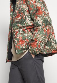 Personne portant une doudoune camouflage verte, rouge et beige avec poches zippées, un pull beige et un pantalon gris foncé, la main dans la poche.