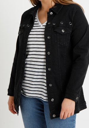 Kvinde iført en sort knappet jakke over en hvid og sortstribet V-hals skjorte og blå jeans.