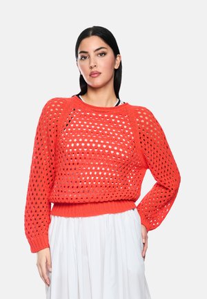 Frau mit langen schwarzen Haaren, die einen leuchtend orangefarbenen offenen Strickpullover über einem weißen Top und einem weißen plissierten Rock trägt, vor einem schlichten Hintergrund stehend.