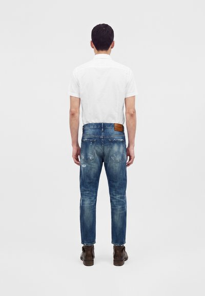 Homme debout de dos, portant une chemise blanche à manches courtes, un jean bleu usé et des bottes en cuir marron sur un fond clair uni.
