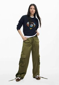 Pull marine avec un motif cœur multicolore, associé à un pantalon cargo vert olive avec poches et taille à cordon. Chaussures marron.