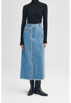 Kvinde iført sort langærmet rullekrave, lyseblå denim midi nederdel med frynsede syninger foran og sorte ankelstøvler på hvid baggrund.