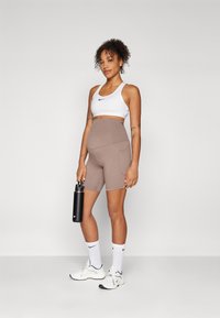 Hvit sports-bh, brune høytlivshorts med lomme, svart vannflaske, hvite sokker med logo, og hvite treningssko. Glatt stoff.