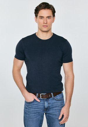 Joven con cabello corto y castaño, vistiendo una camiseta ajustada azul marino, jeans azules y un cinturón marrón, de pie con una mano en el bolsillo.