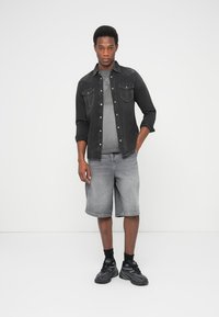 Camicia di denim nera, t-shirt grigia con grafica e pantaloncini grigi; modello che indossa scarpe da ginnastica nere con suola testurizzata; sfondo neutro.