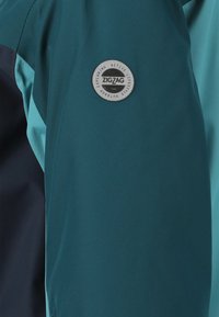 Tyrkis- og marineblå jakke med en glat tekstur, der har et rundt metal-logo på venstre ærme. Designet inkluderer paneler i kontrastfarver.