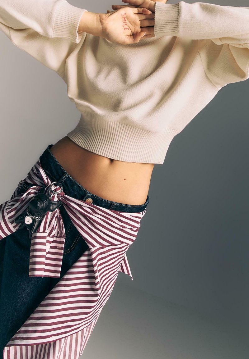 Personne portant un pull crème avec une chemise rayée nouée autour d'un jean foncé, mains couvrant le visage, le mot "SOURIRE" écrit sur la paume gauche.