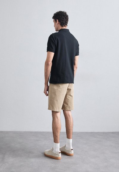 Melns polo krekls, bēšas šortas, kedas ar gumijas zoli. Modelis vērsts prom, rādot īsās piedurknes un minimālistisku dizainu.