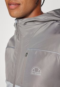 Svijetlosiva jakna protiv vjetra s patentnim zatvaračem sprijeda, reflektirajućim detaljima i Ellesse logotipom. Glatka tekstura s prilagodljivom kapuljačom.