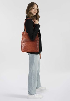 Bruine leren tote bag met een zachte textuur, uitgerust met een ritszak en lange handvatten, gedragen door een model met lichte jeans en een zwarte jas.