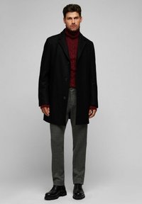 Abrigo negro, sudadera de cuello alto burdeos texturizada, pantalones de cuadros grises y zapatos negros. Presenta botones estándar y un ajuste a medida.